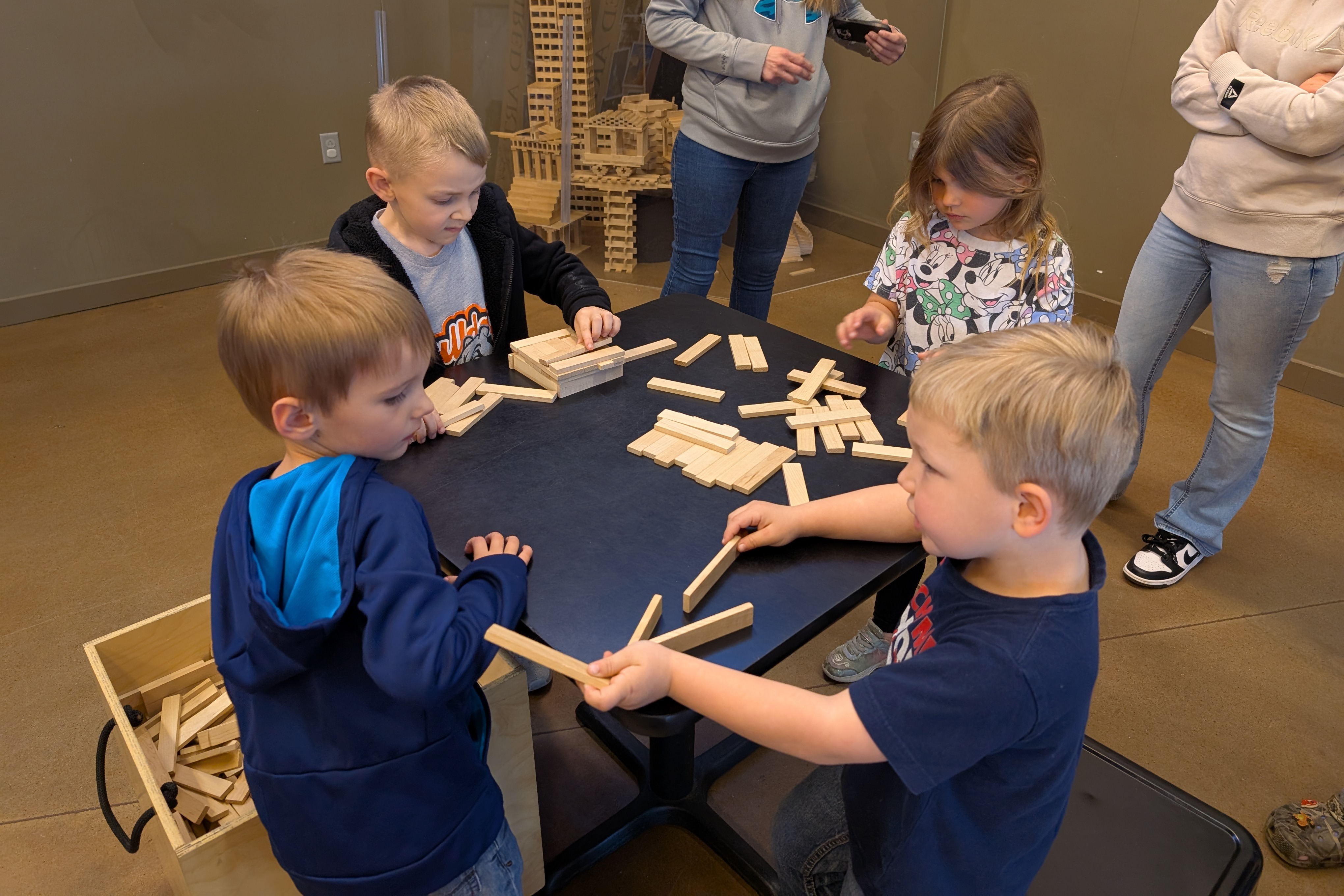 A group of children play with blocks at a table in a room, with adults standing nearby.
