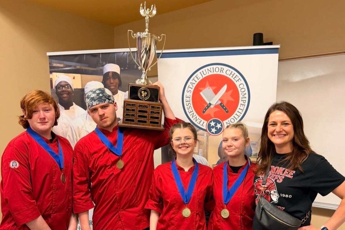 Five people in red chef uniforms stand by a banner, one holding a trophy. They wear medals.