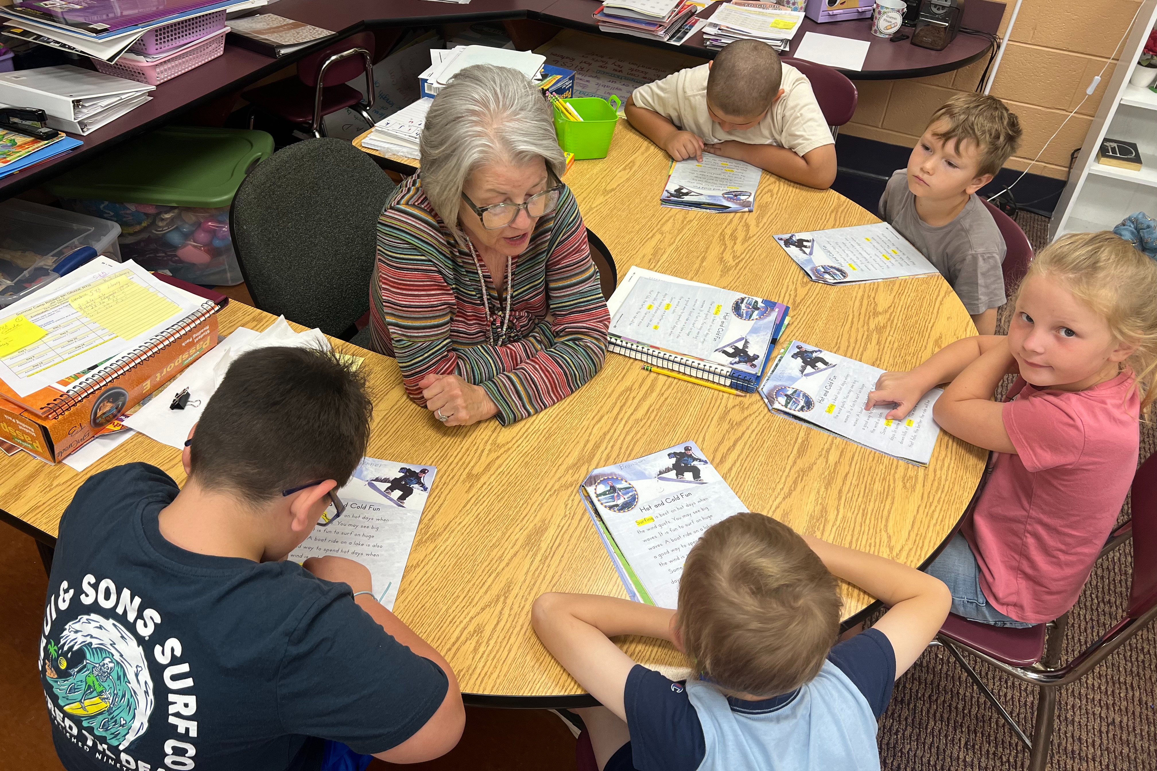 A teacher with glasses sits at a table with five children. They all have books in front of them.