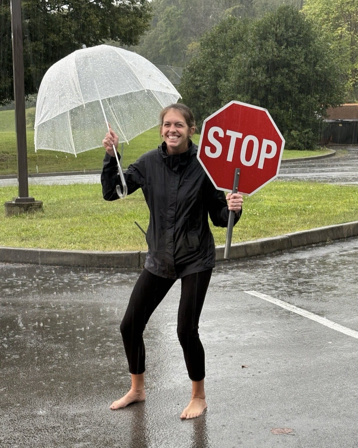 Woman standing in wet road holding clear umbrella and red stop sign. Trees and grass in background.