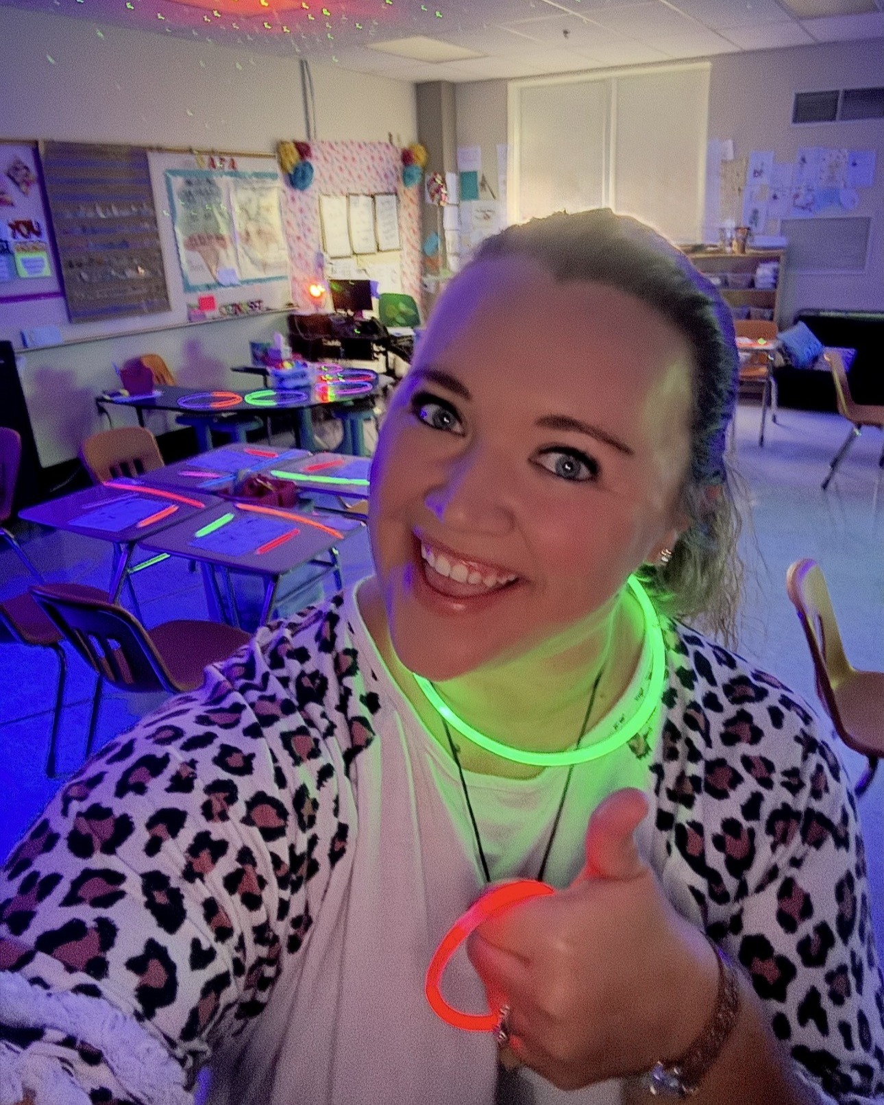 A smiling woman wearing neon green accessories stands in a classroom with desks and chairs.