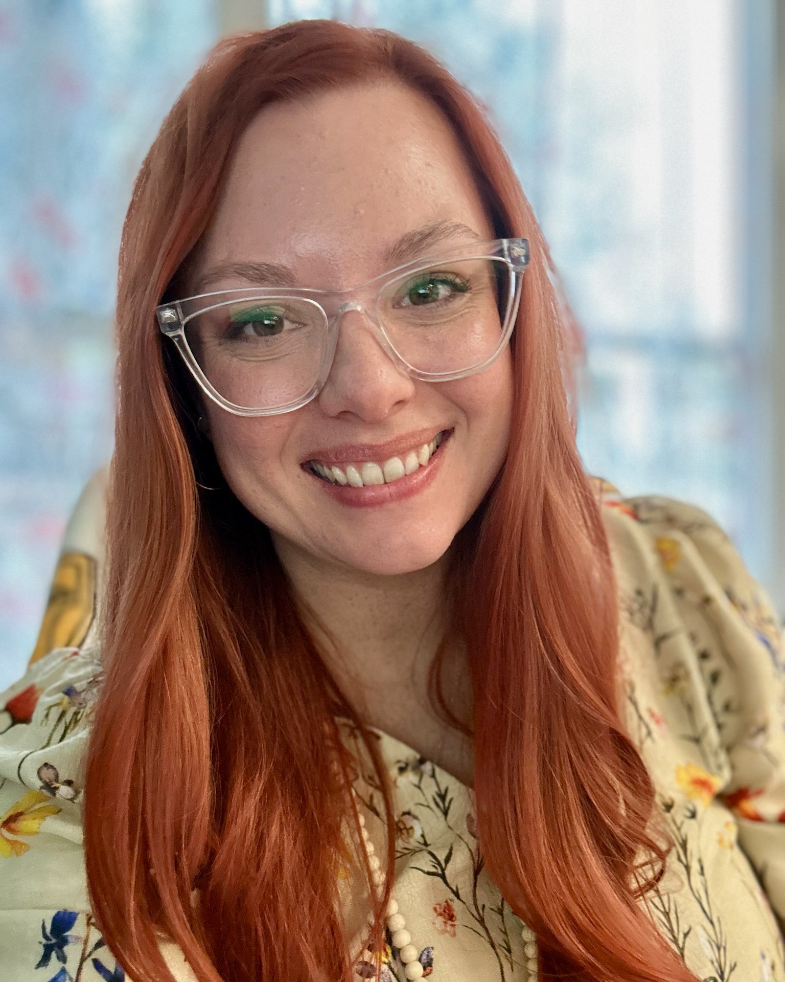 Woman with long red hair wearing clear glasses and a floral-patterned blouse, smiling at the camera.