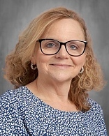 A woman with blonde, curly hair and glasses smiles. She wears a blue and white patterned blouse.
