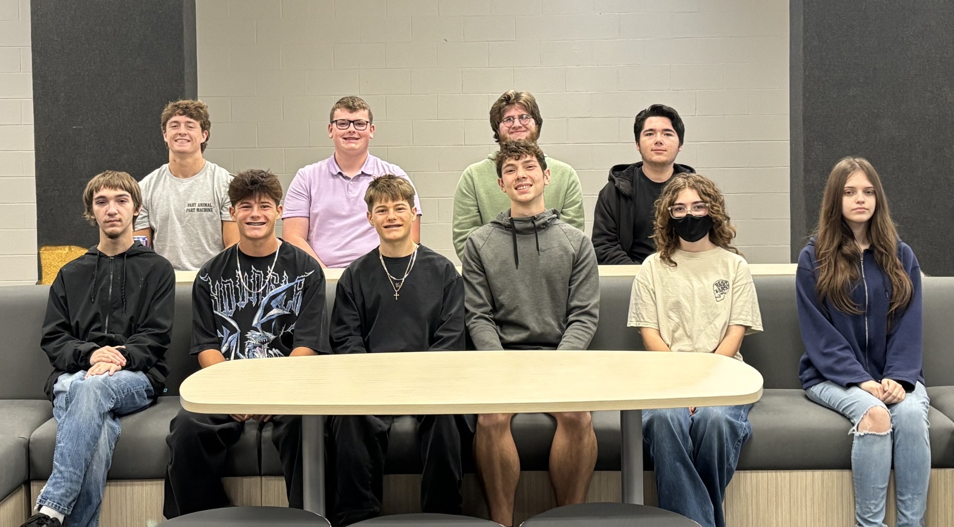 A group of teens wearing casual clothing sit around a table in a room with white walls.