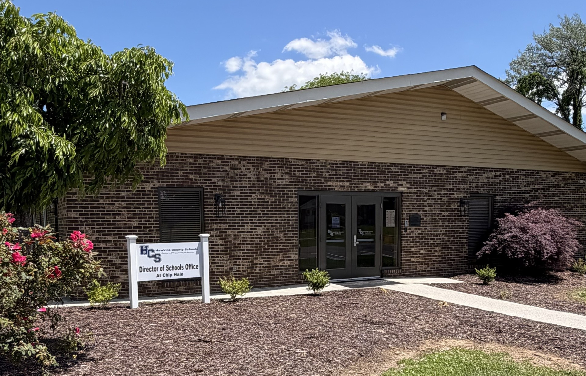 A brick building with glass doors and windows stands with a sign. A white sign with text stands in front.