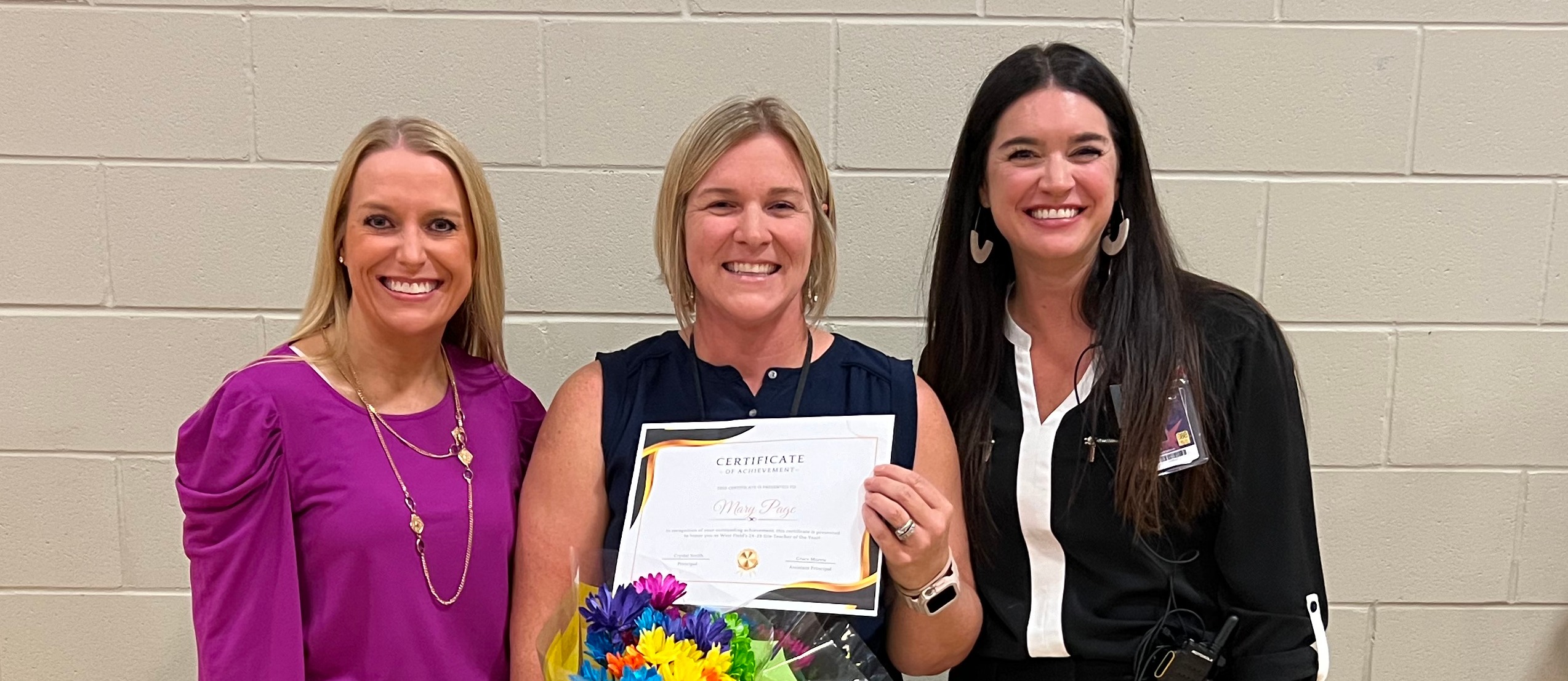 Principal, assistant principal and teacher of the year Mary Page standing in gym holding flowers