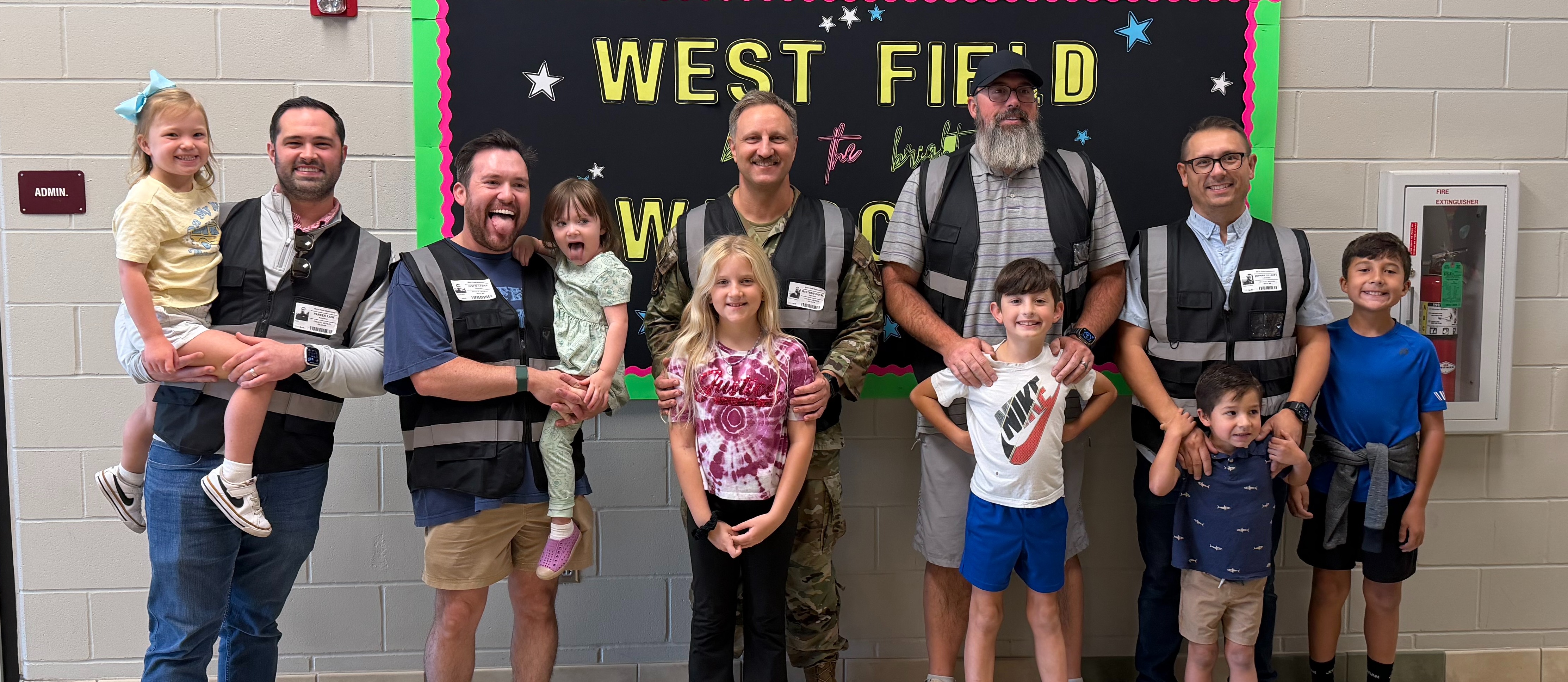 A group of smiling adults and children stand in front of a school welcome sign. The kids appear happy and are being held or standing beside the adults.