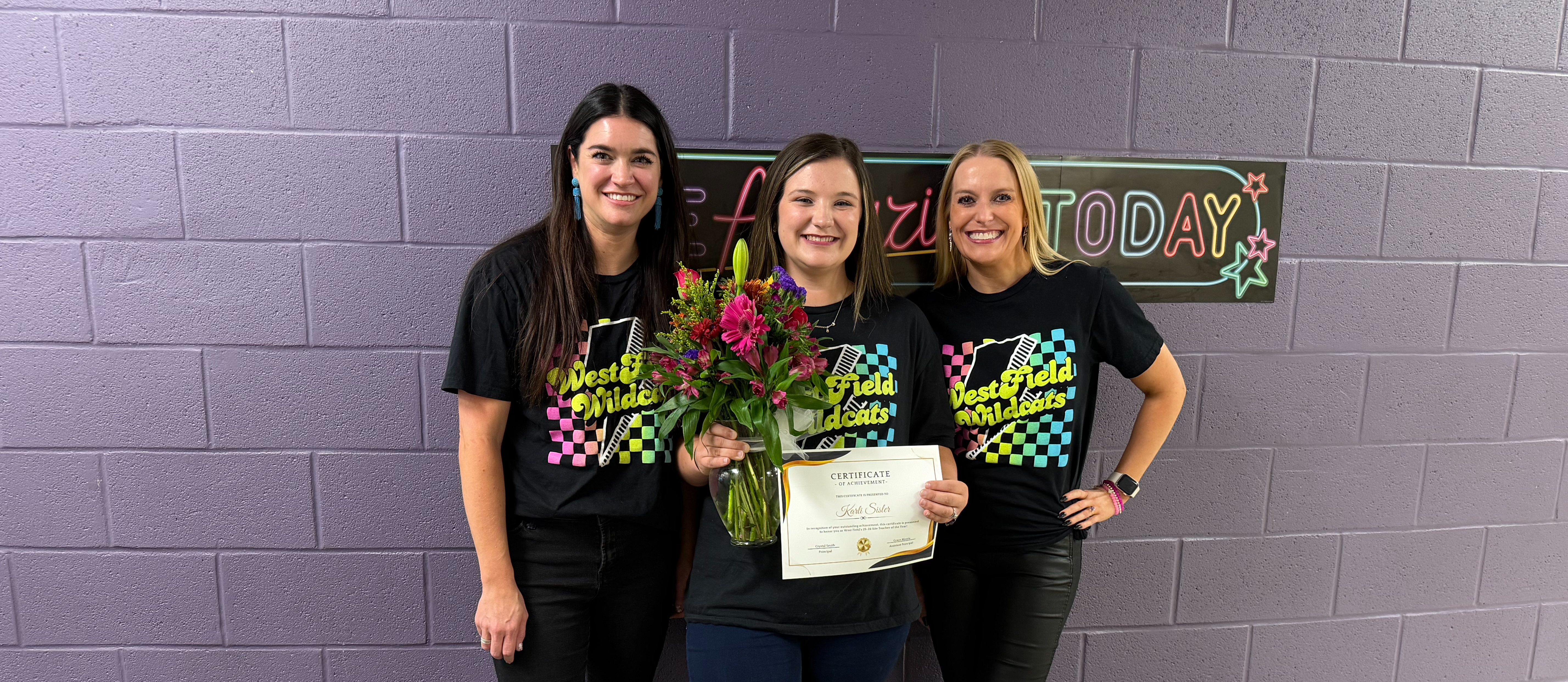 Three women smiling in front of a purple wall. The center holds flowers and a certificate. All wear matching "Westfield Wildcats" shirts. Festive atmosphere.