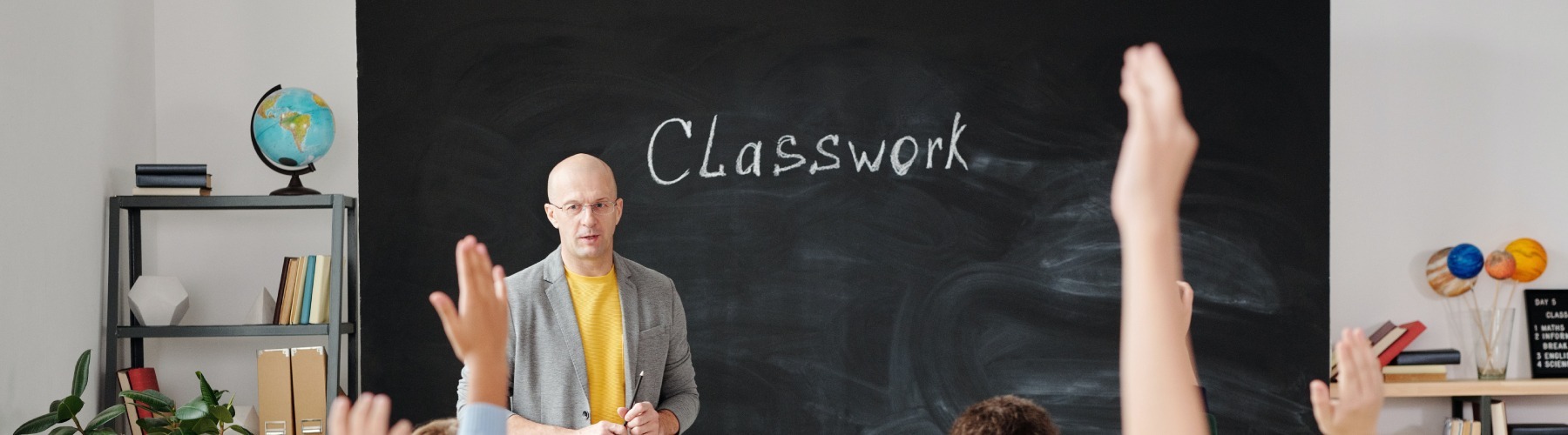a teacher stands in front of a blackboard that reads "classwork" while several students raise their hand to ask or answer a question