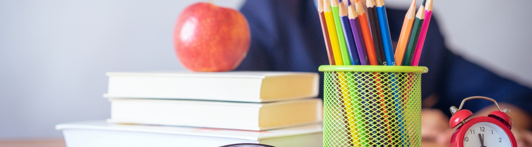an apple on a stack of books next to a cup of pencils and an alarm clock. a student is in the background