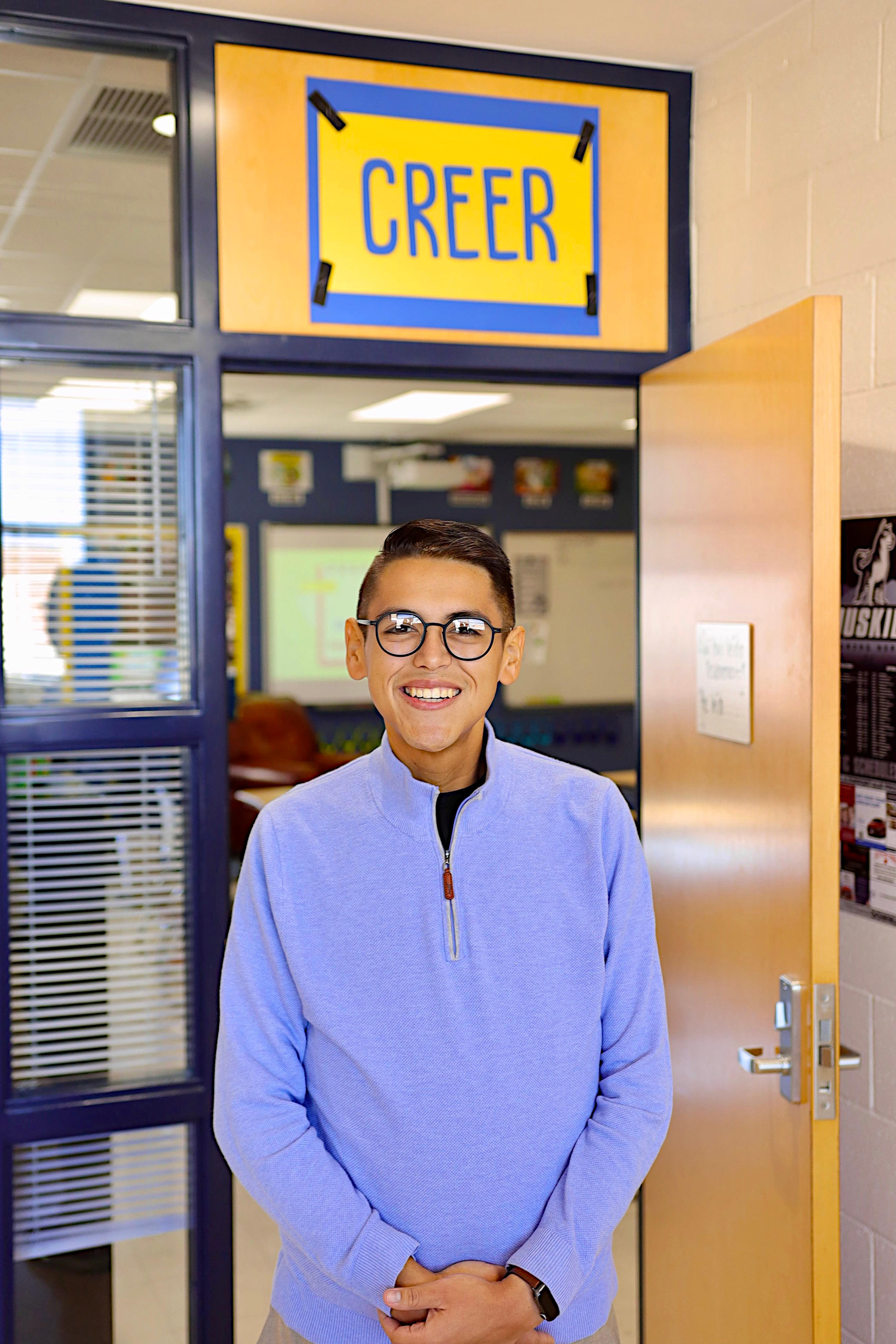 Ezequiel Madrigal, Teacher of the Year, is depicted as a young man in a blue sweater and glasses.