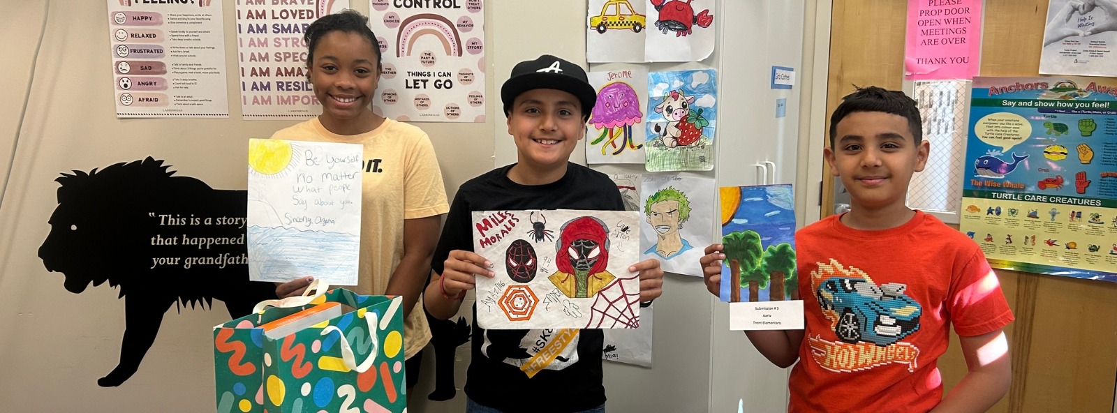 Three kids in a classroom holding drawings. A black bear drawing is on the wall behind them.