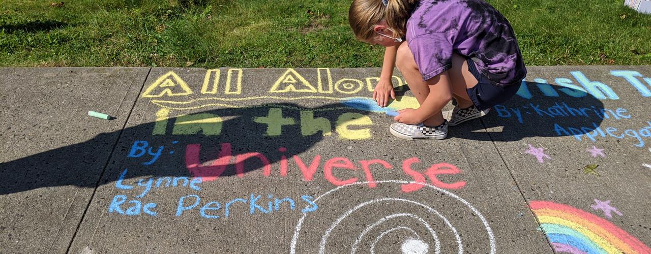A close up of a student drawing the title "all alone in the universe" book title with chalk drawing next to it.