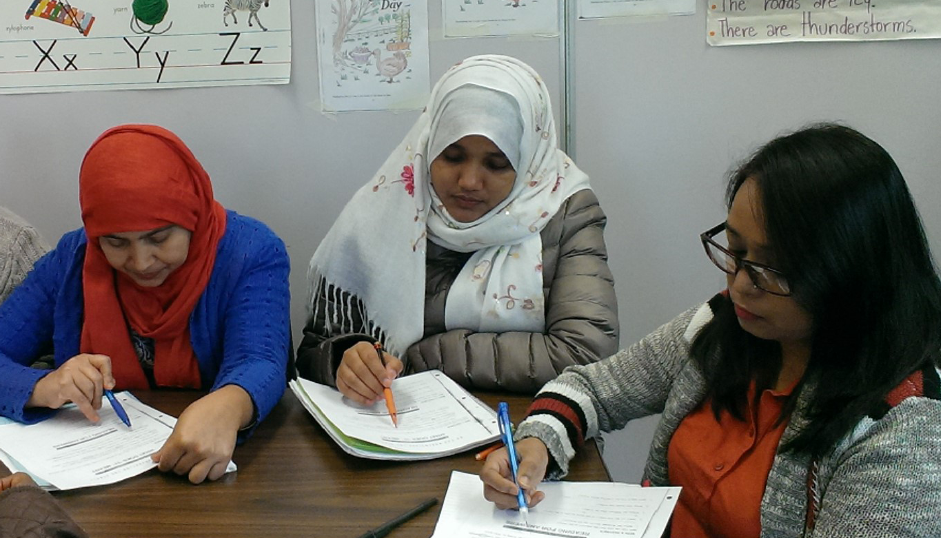 Three adult ESL classmates writing in notebooks at desk