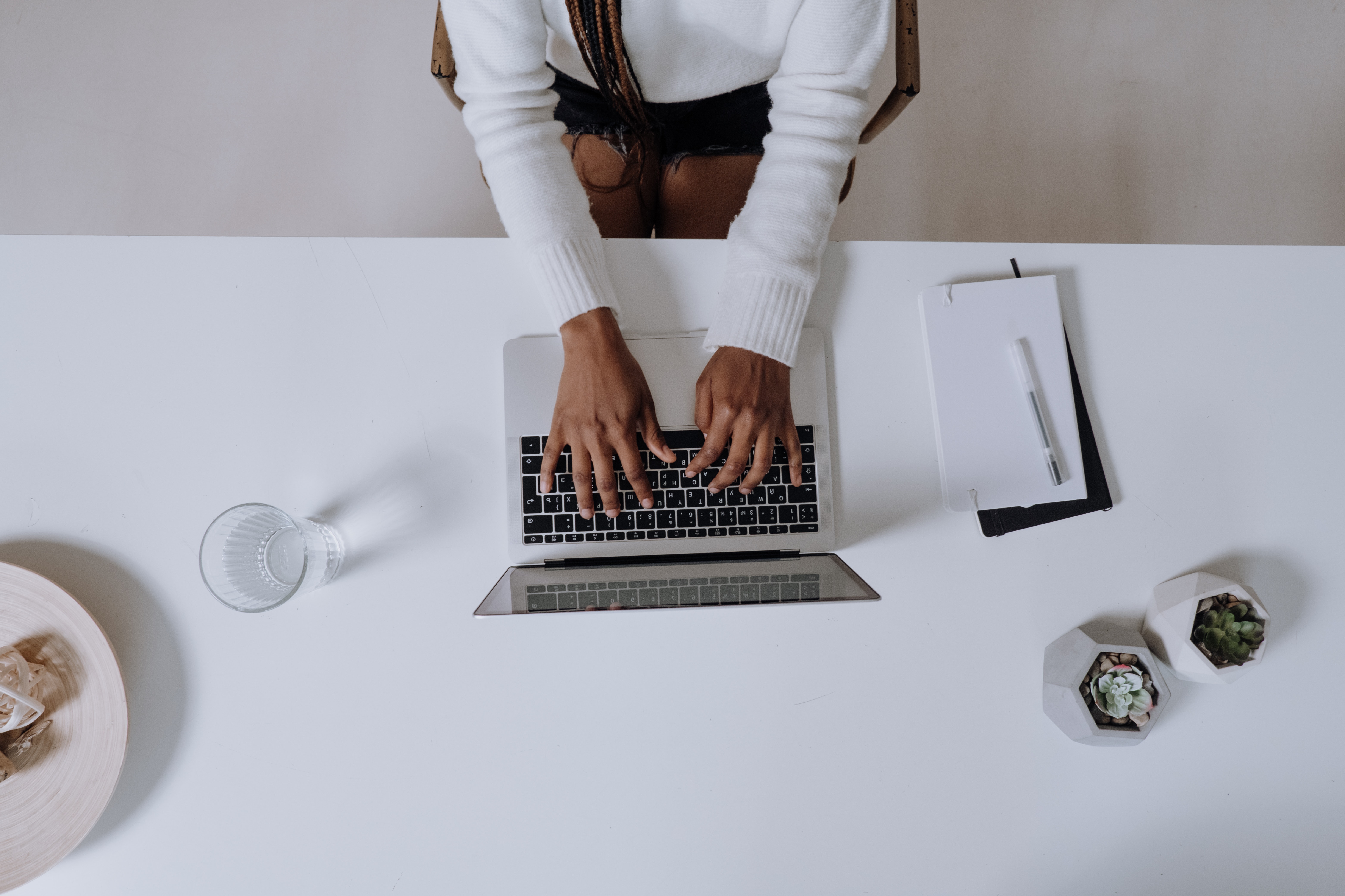 A bird's eye shot of an individual typing on a laptop. The laptop sits on a white desk with cords on either side. 