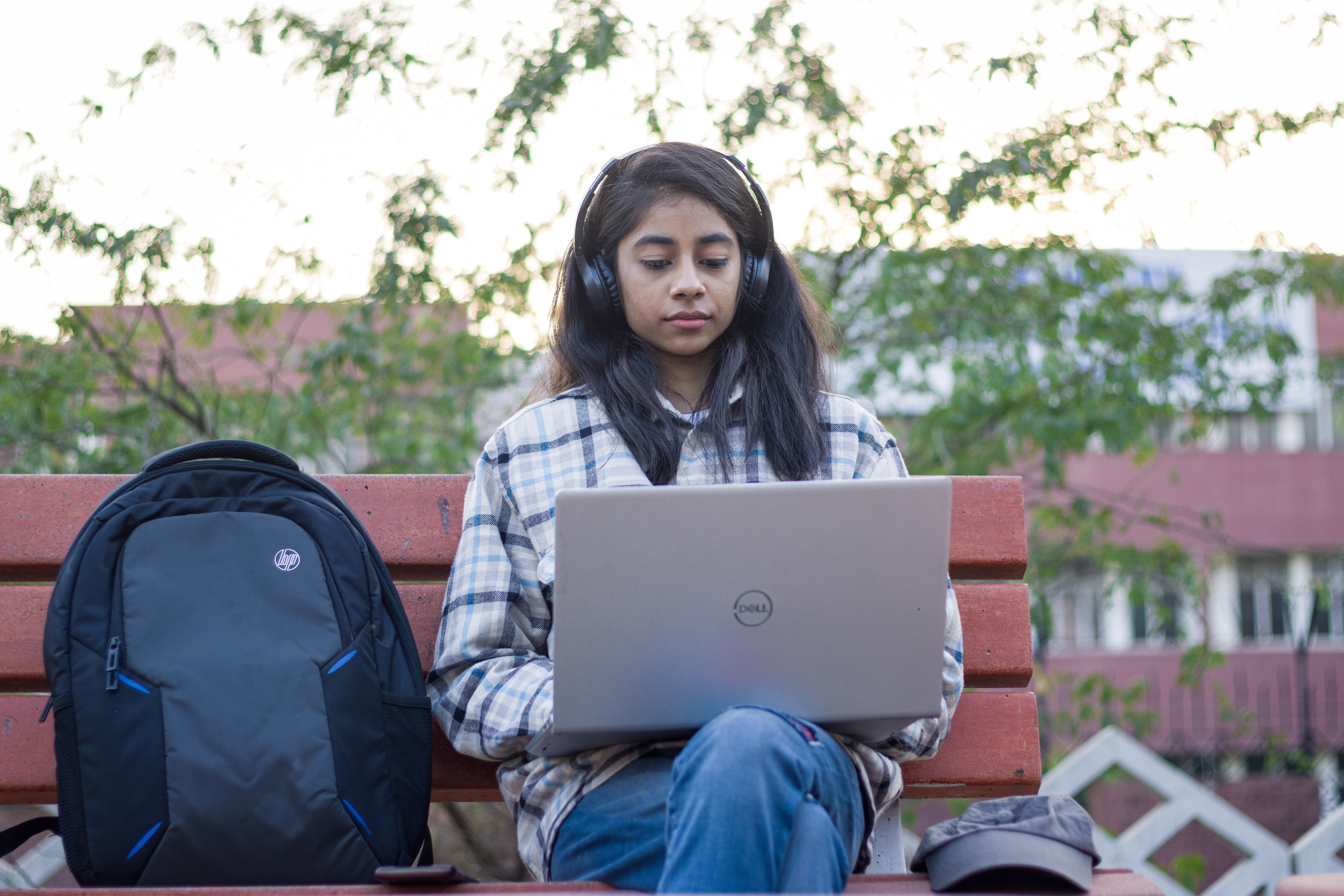 Student sits on a bench working on her laptop. Her backpack sits beside her and she wears headphones while she works. 