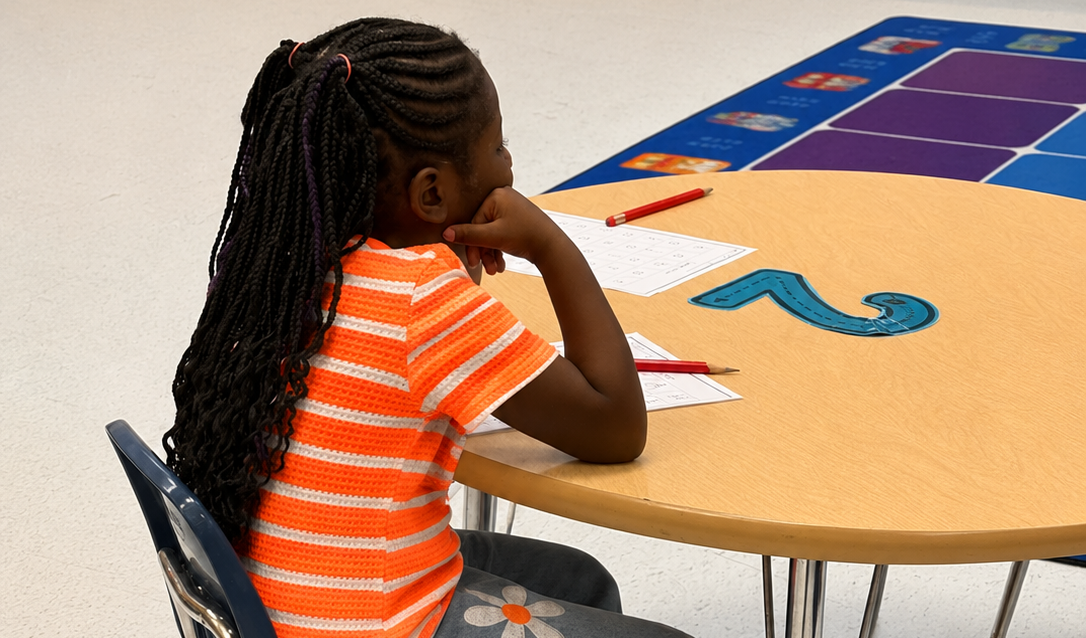 A student sits at a classroom table completing a worksheet with pencils and learning materials nearby. The student is focused on the activity in a colorful elementary classroom setting.