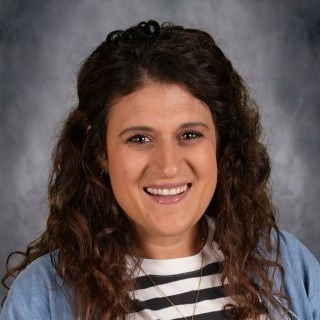 A woman with dark curly hair smiles. She wears a white and black striped shirt and a blue cardigan.