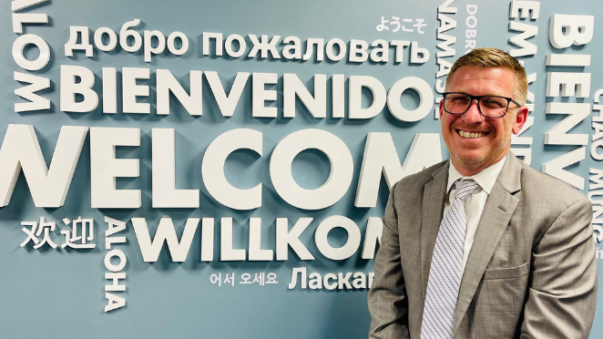 A man in a business suit and glasses stands in front of a blue wall with various languages on it.