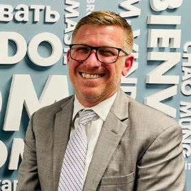 Man in business attire stands before a wall with multiple languages saying "welcome", "bienvenido", "willkommen", etc.