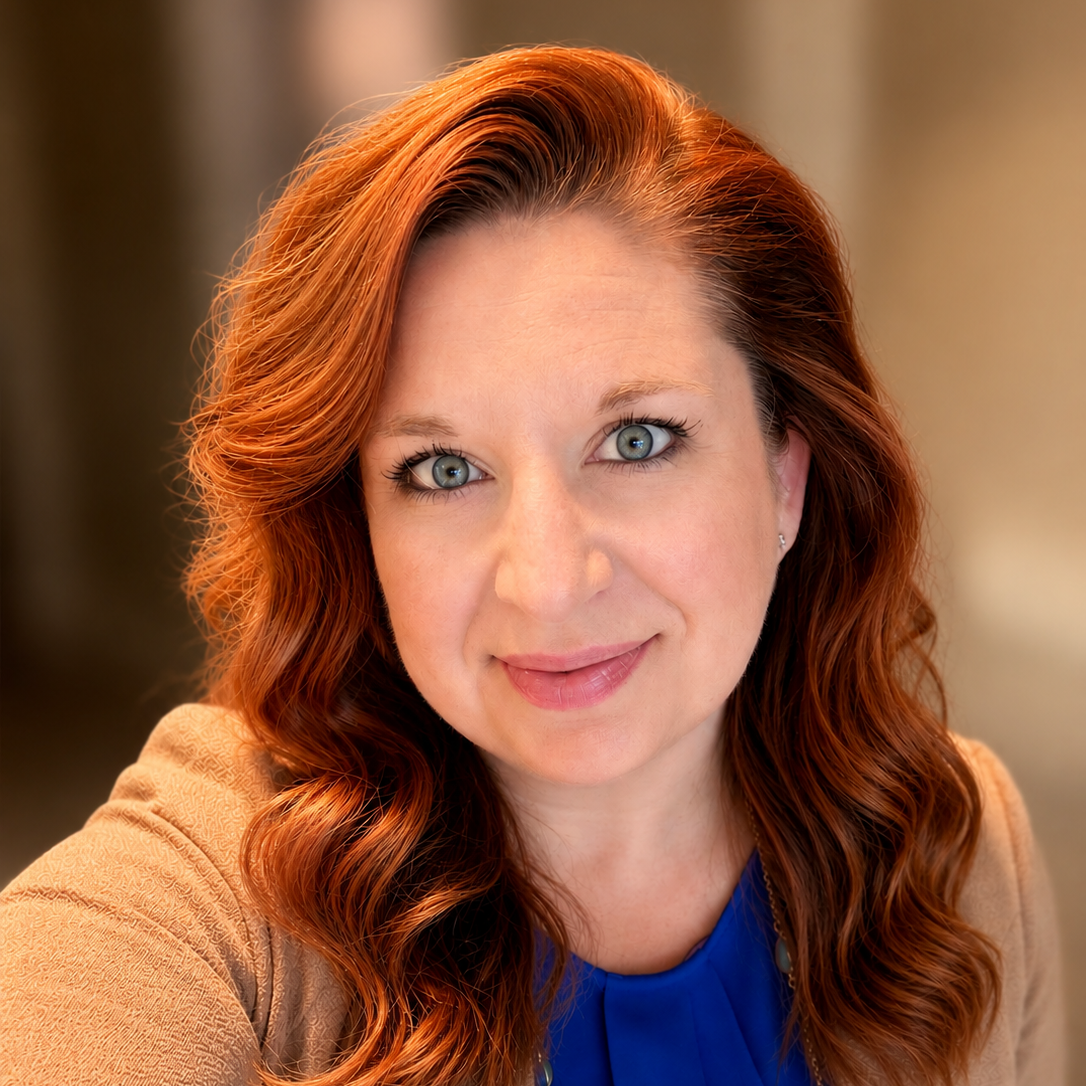 A woman with red curly hair and blue eyes wearing a blue top and a light brown jacket.