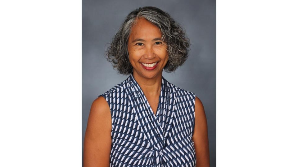 A woman with gray hair smiles, wearing a blue and white patterned top. The background is a plain gray.