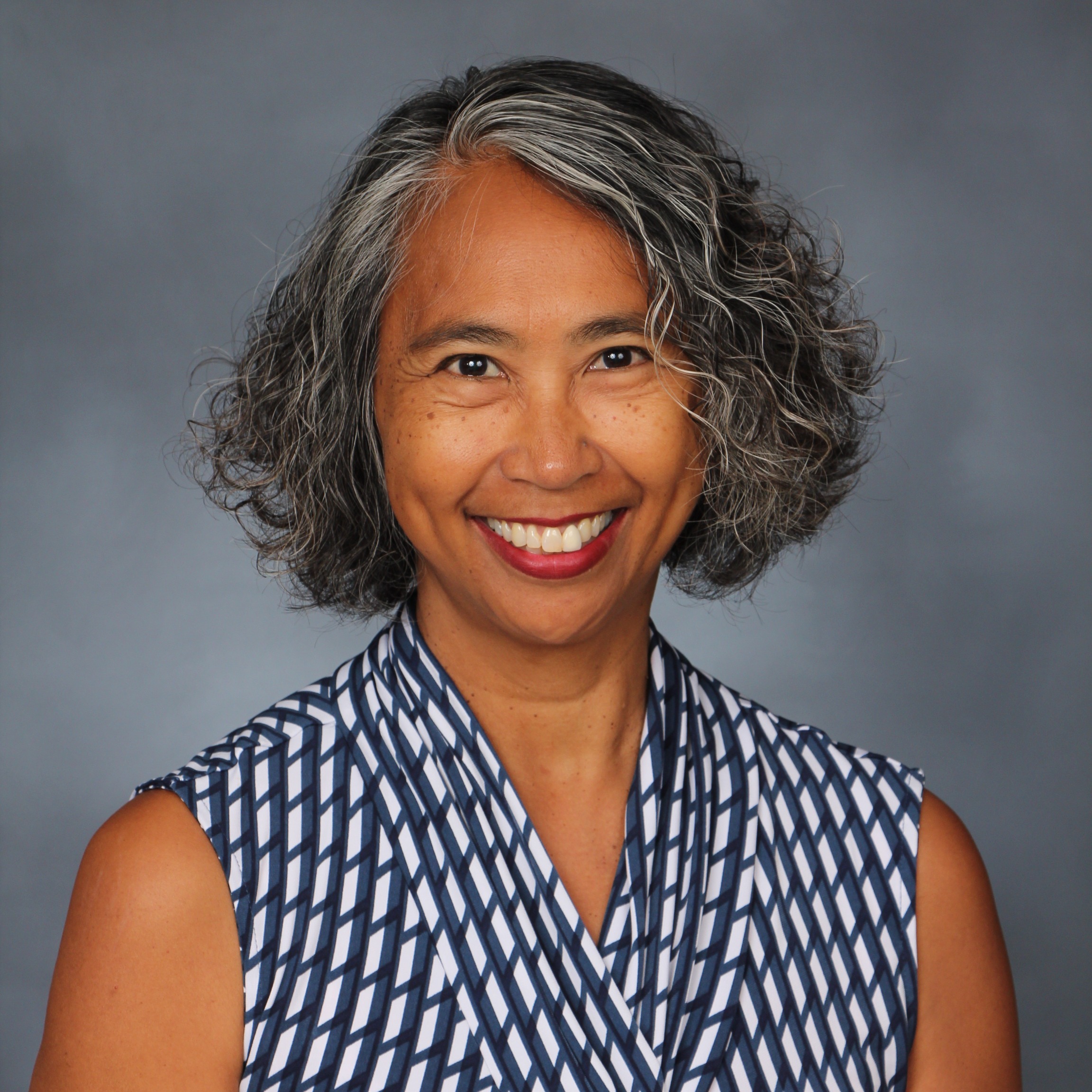 A woman with short, gray hair and a blue-and-white patterned top smiles, slightly raising her arms.