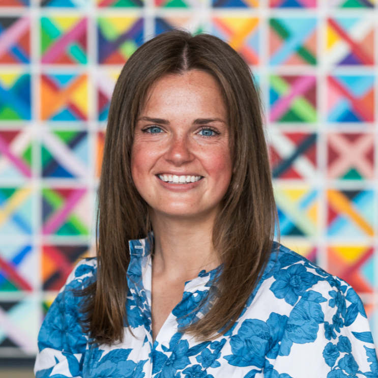 Woman with blue eyes and brown, straight hair, wearing blue floral shirt against a colorful geometric background