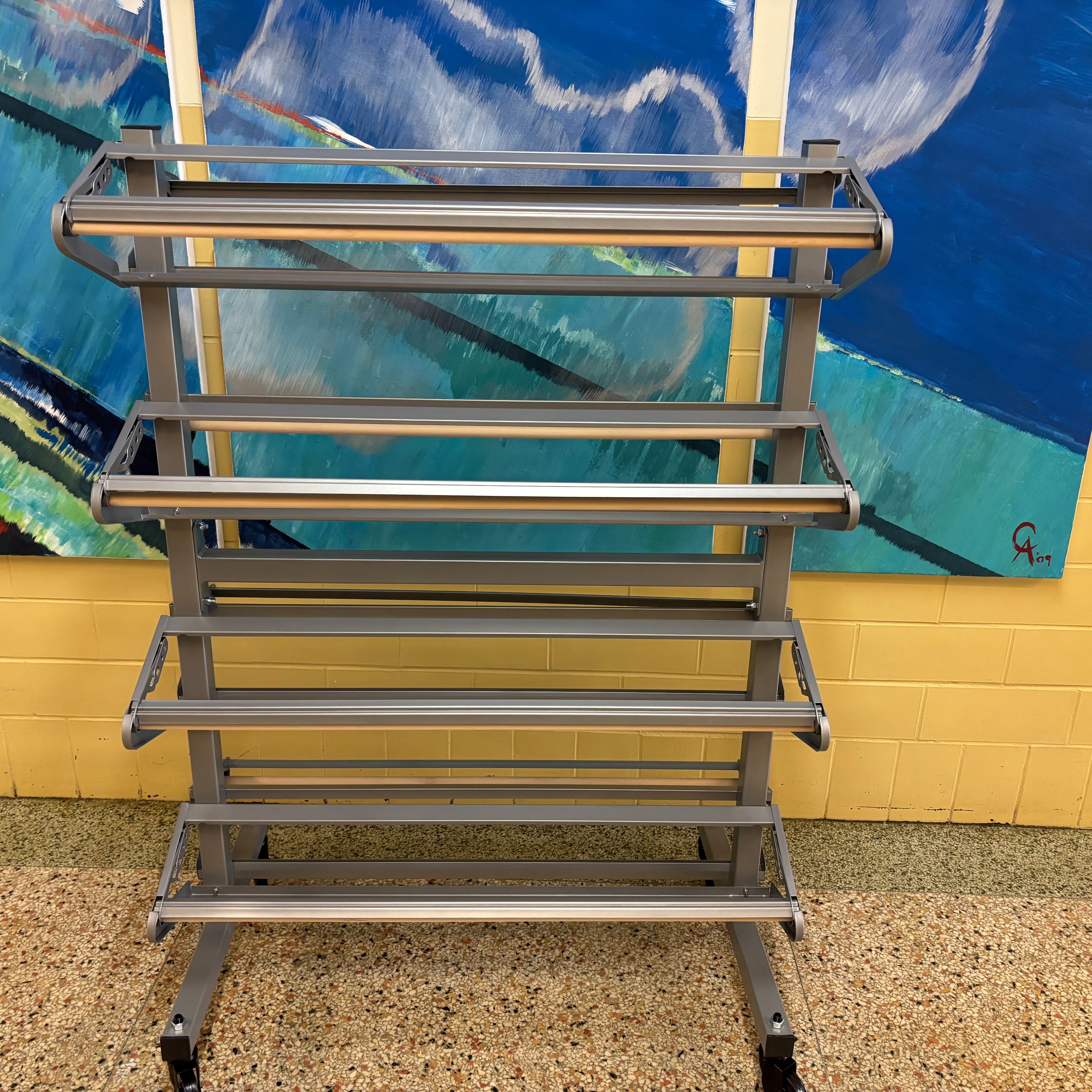 A rolling cart with three shelves stands before a yellow wall and two blue paintings.