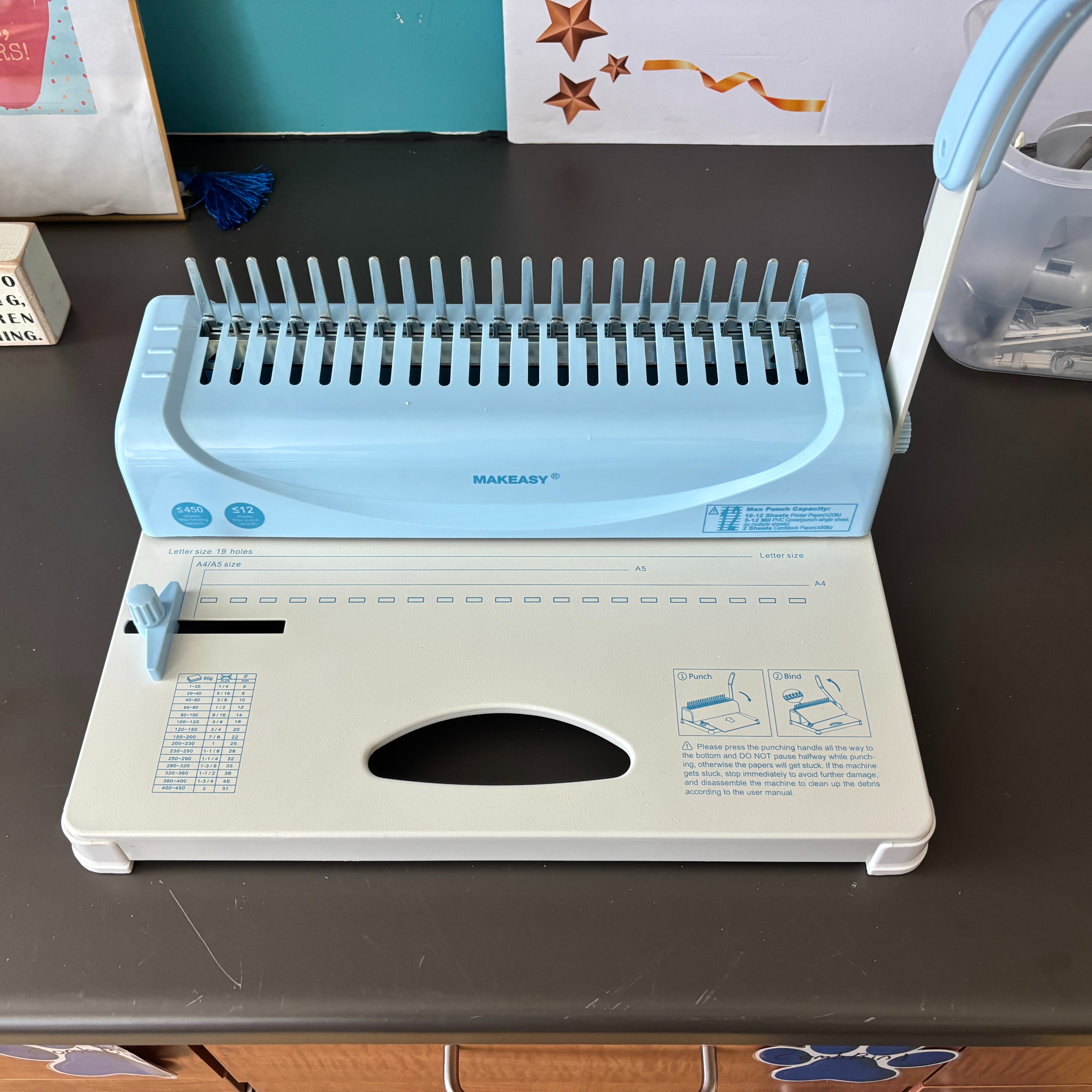 A blue and white hole punch sits on a brown table with a plastic container on the side.