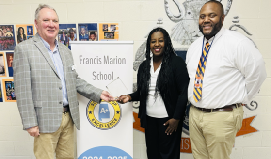 Three adults stand indoors in a school hallway next to a vertical sign that reads “Francis Marion School,” features an A+ College Ready seal, and lists “2024–2025.” The person on the left wears a blazer and holds a certificate or document, the person in the center wears a black suit and smiles, and the person on the right wears a white shirt with a striped tie. A school mural and photo display are visible in the background.