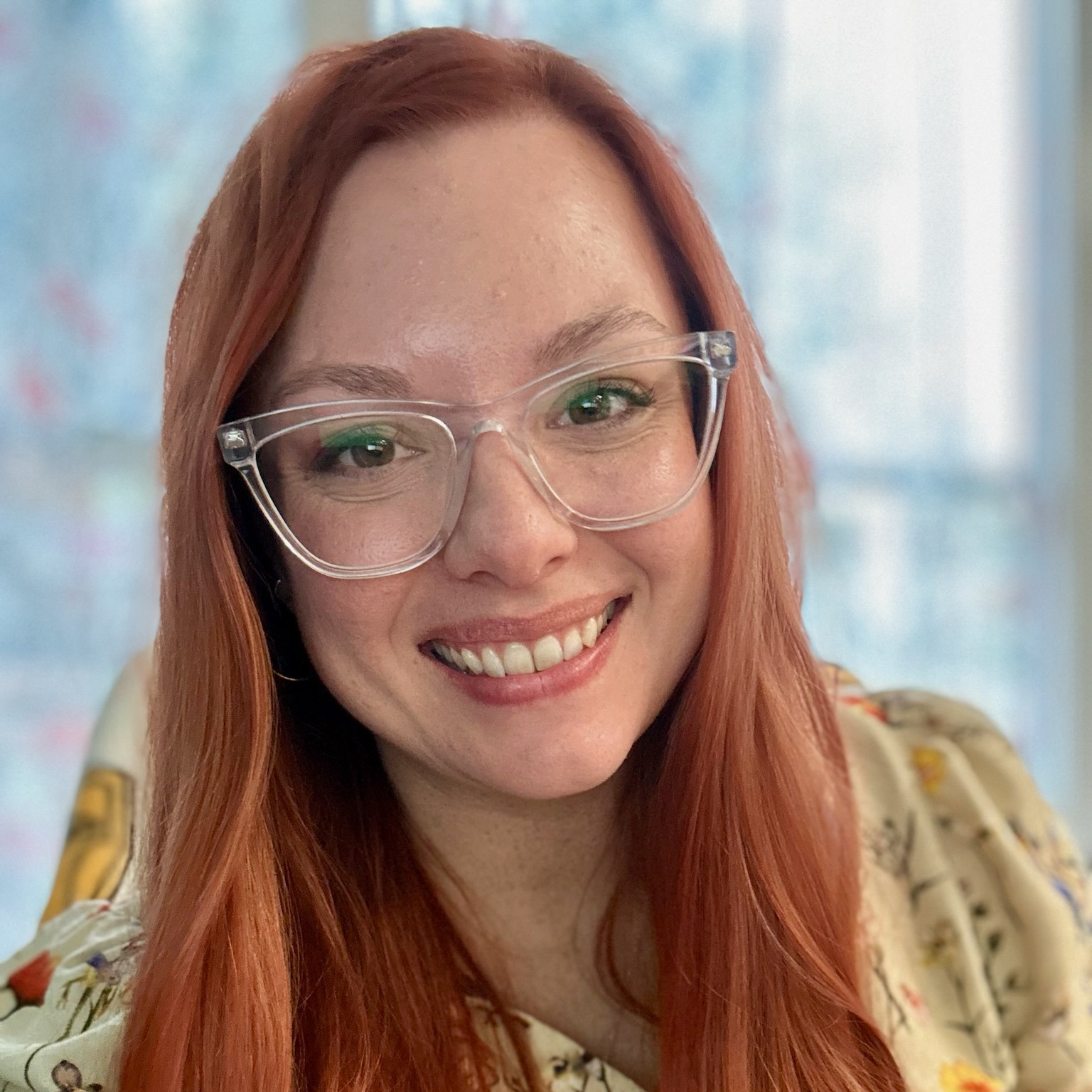 Woman with red hair wearing glasses and a floral blouse, smiling for a close-up portrait.