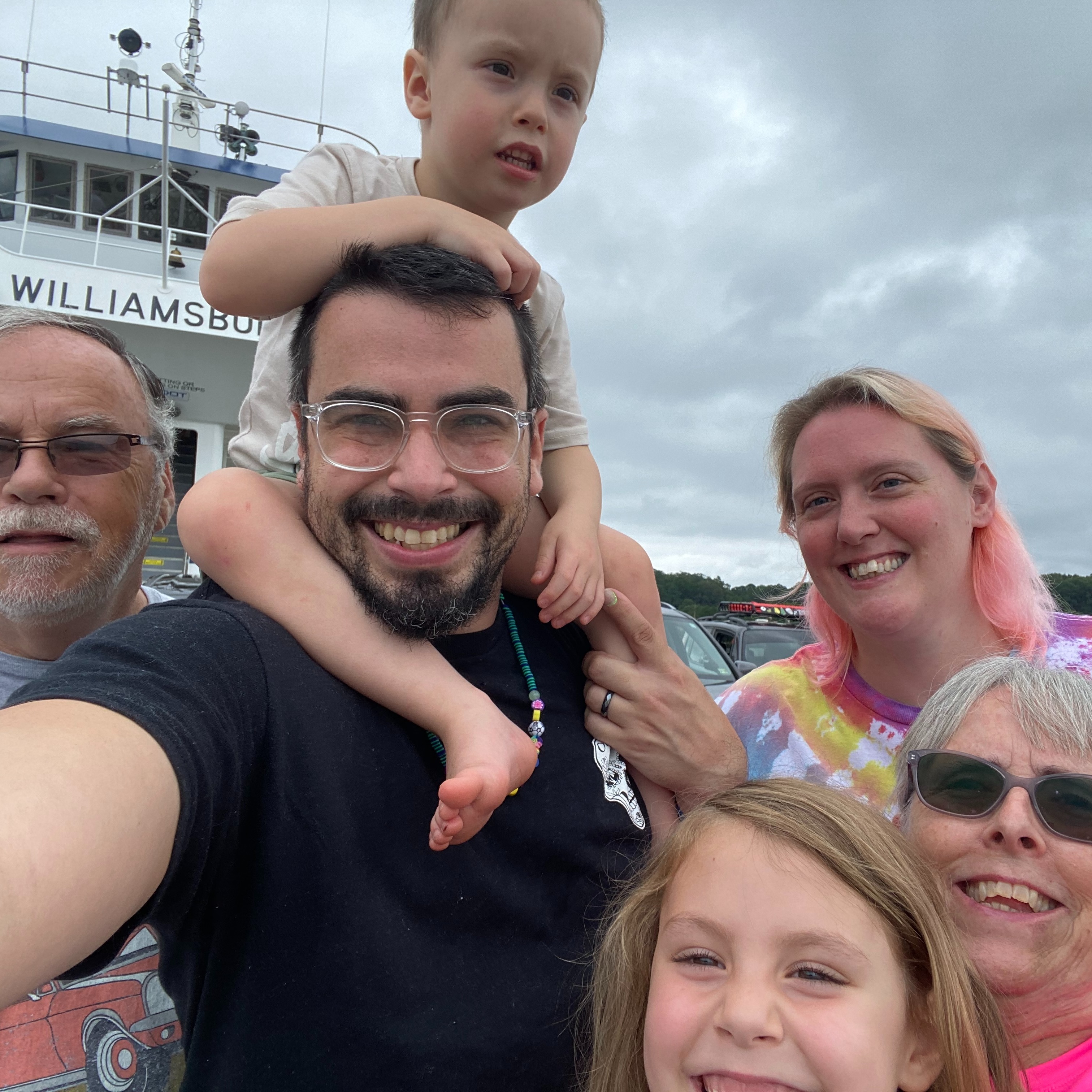A family of five people smiles in a photo, with a child on a man's shoulders, against a cloudy sky.