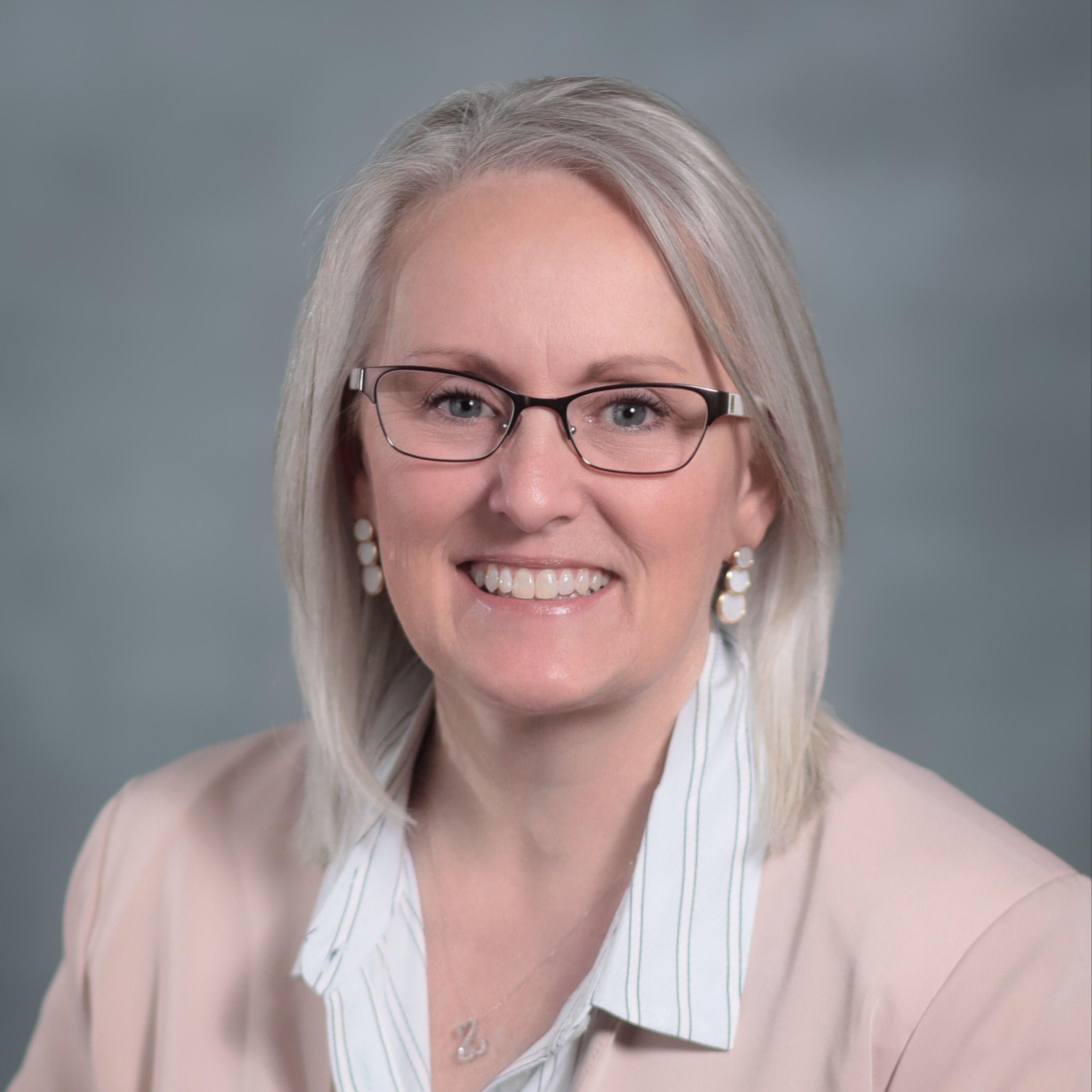 A woman with light hair wears glasses, a necklace, and a light pink blazer. She smiles.