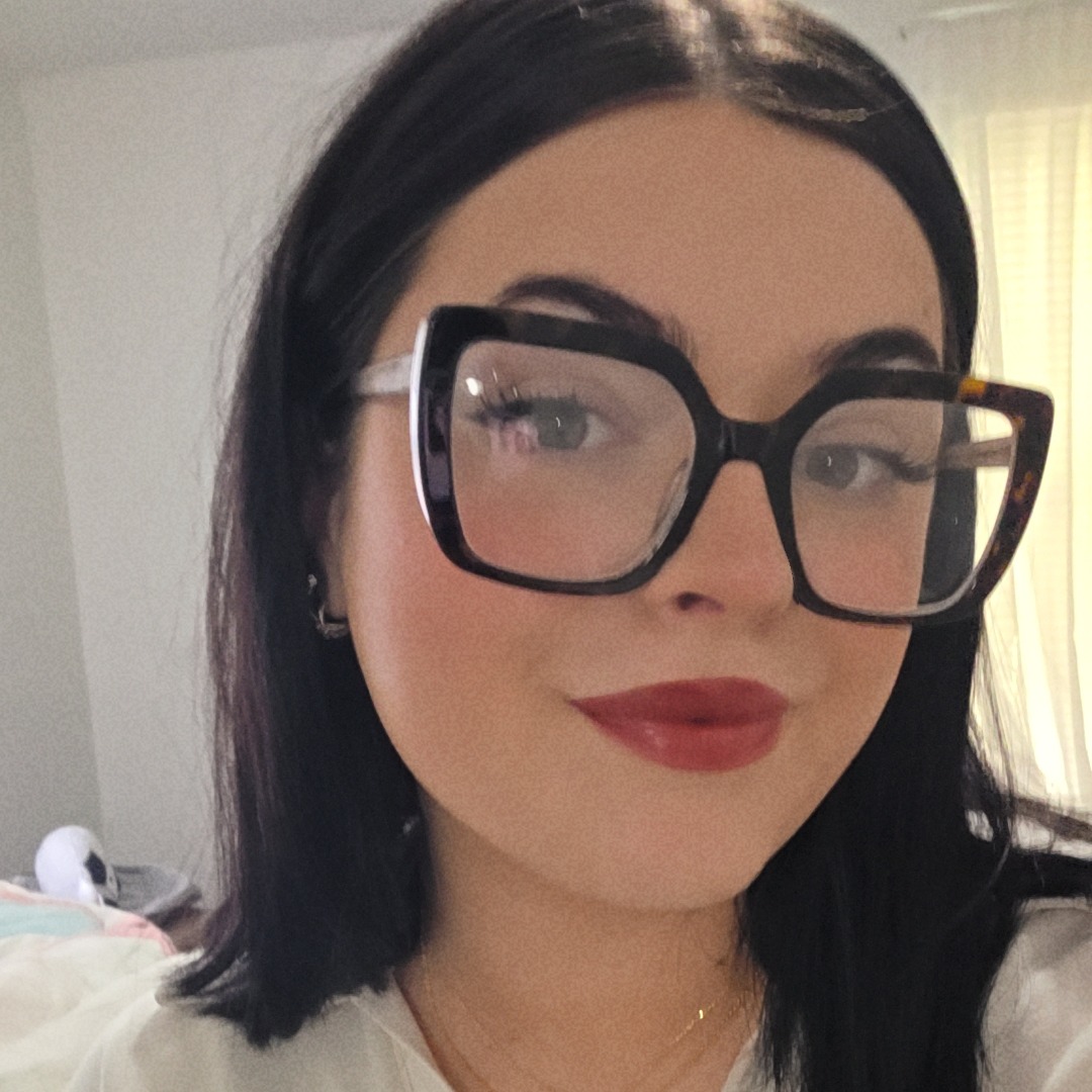 A woman wearing glasses and a white V-neck shirt. She has dark hair, red lipstick, and is in a room.