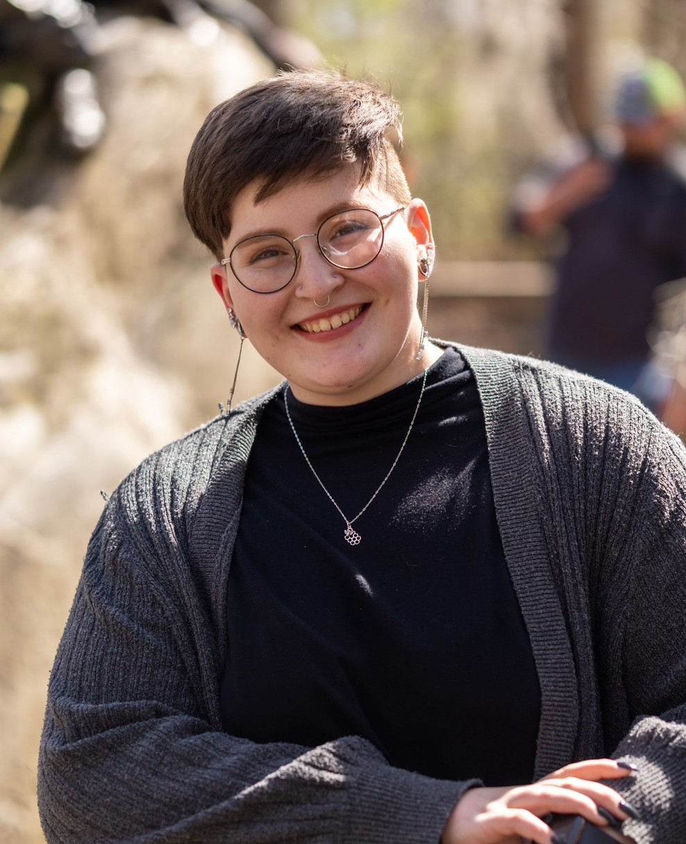 a photo of Mrs. Artemis - they have short brown hair and round glasses. they are smiling and wearing a black shirt with a gray cardigan.