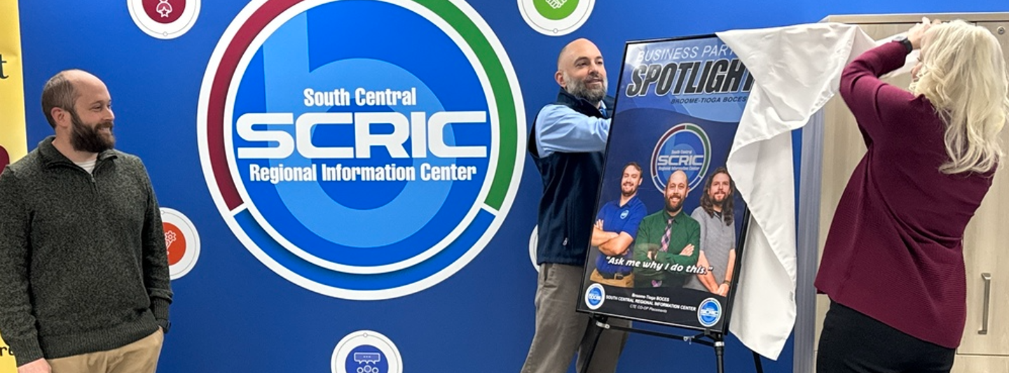 Business Spotlight Award. Two people unleash a bulletin of the Business Spotlight Award by pulling a sheet off that was covering it. Phil Phata who awaits the reveal stands with a smile. 