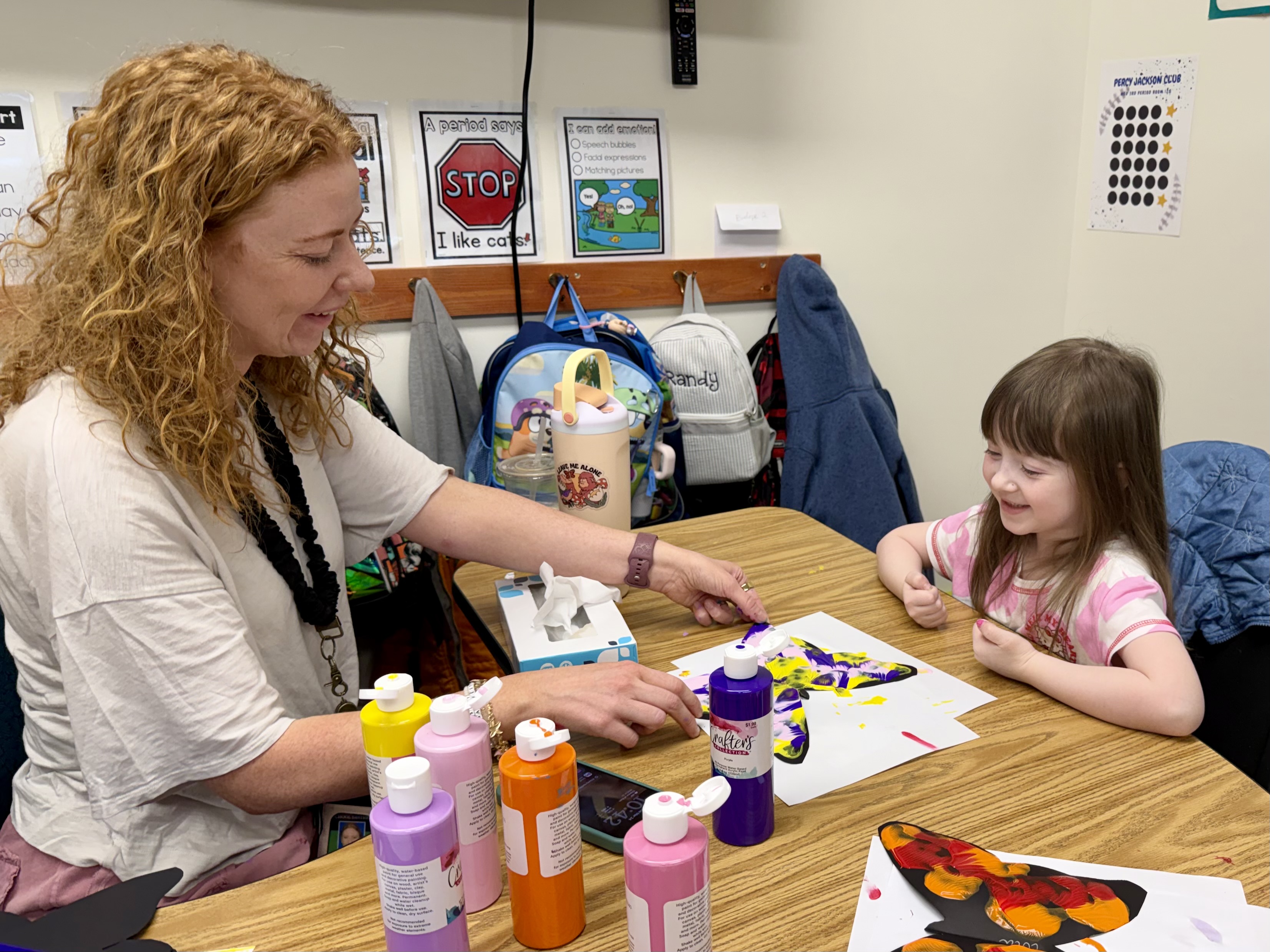 Woman and girl at a table with a butterfly art project. Bottles of paint and a backpack in the background.