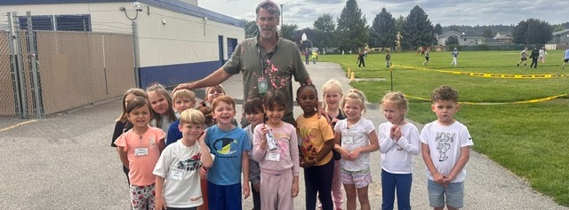 A group of young children stands with an adult in front of a school building. The background shows a grassy field with a fence.