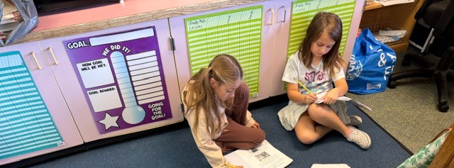 Four children sit on the floor, engrossed in reading books. A carpeted area, school furniture, and a trash bin are visible.