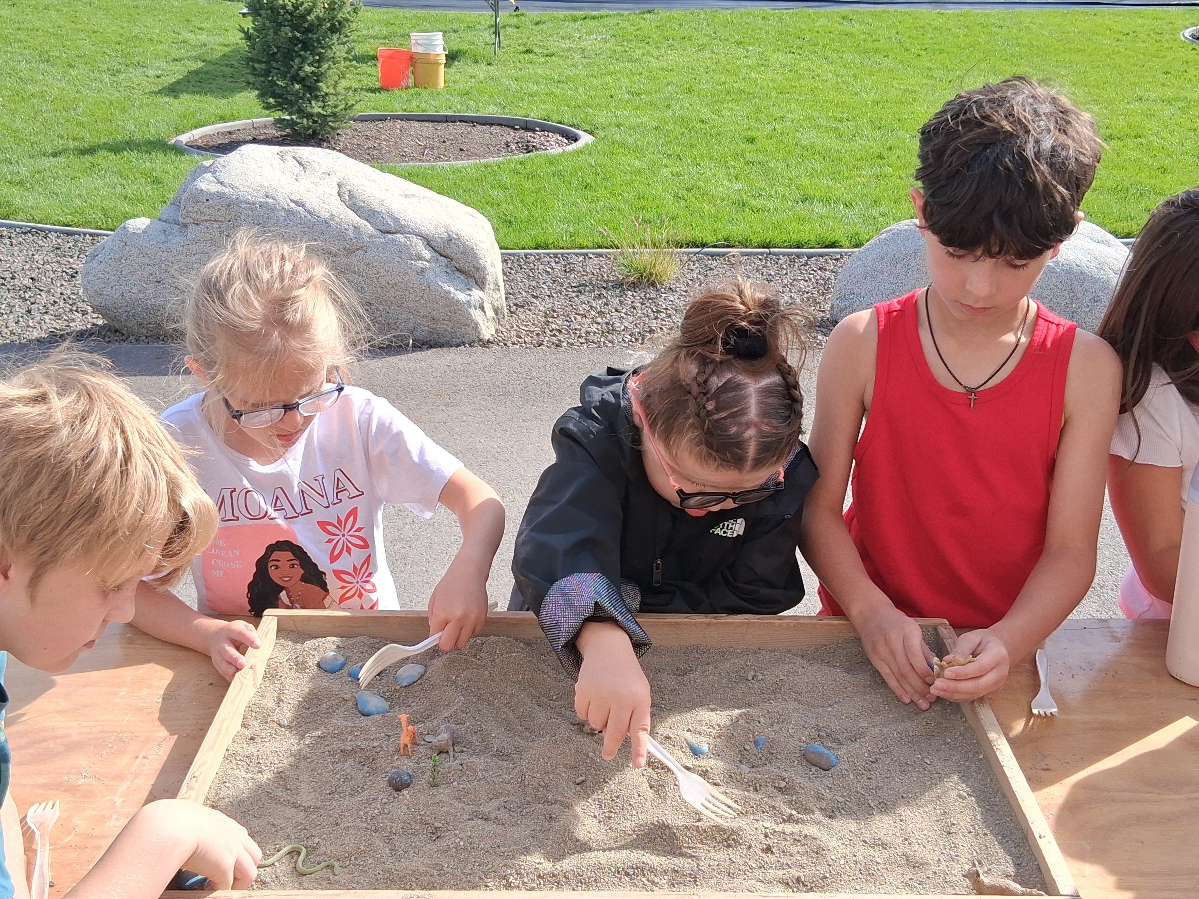 Several children are playing in a sandbox with small tools, set on a concrete surface. A grassy area is in the background.