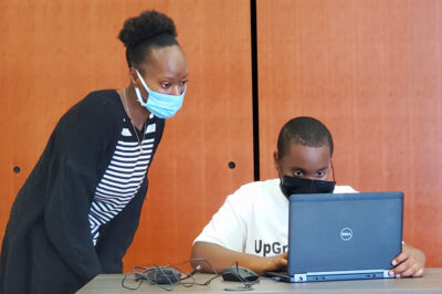 teacher wearing mask looks over shoulder of student on a computer