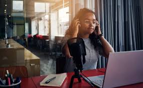 student wearing headphones sits in room with lots of natural light in the background in front of a laptop and microphone 