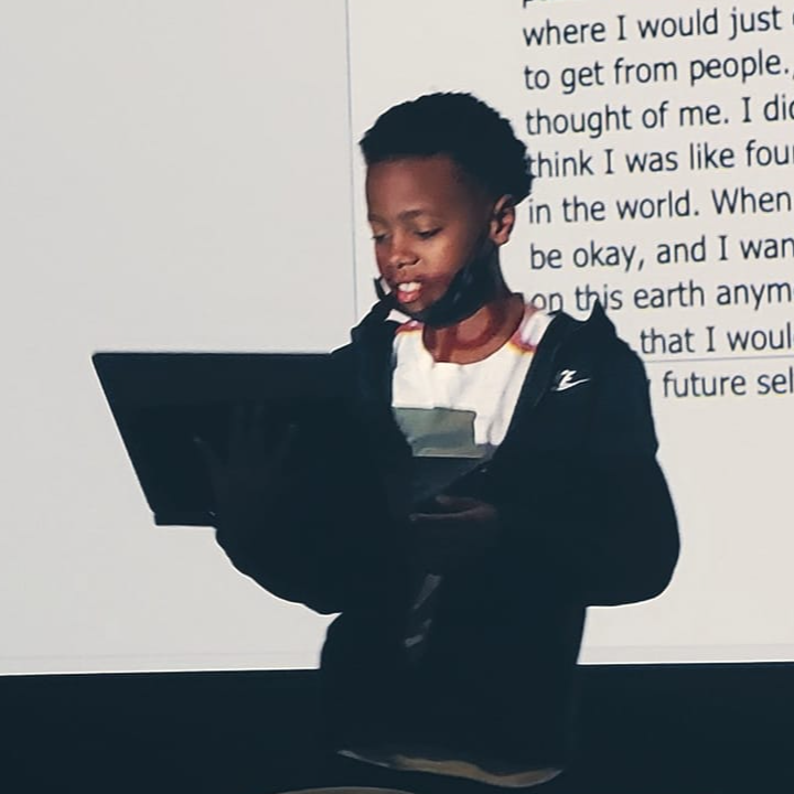 Jovan holding a laptop standing on stage reading "Younger Self," which is projected behind him