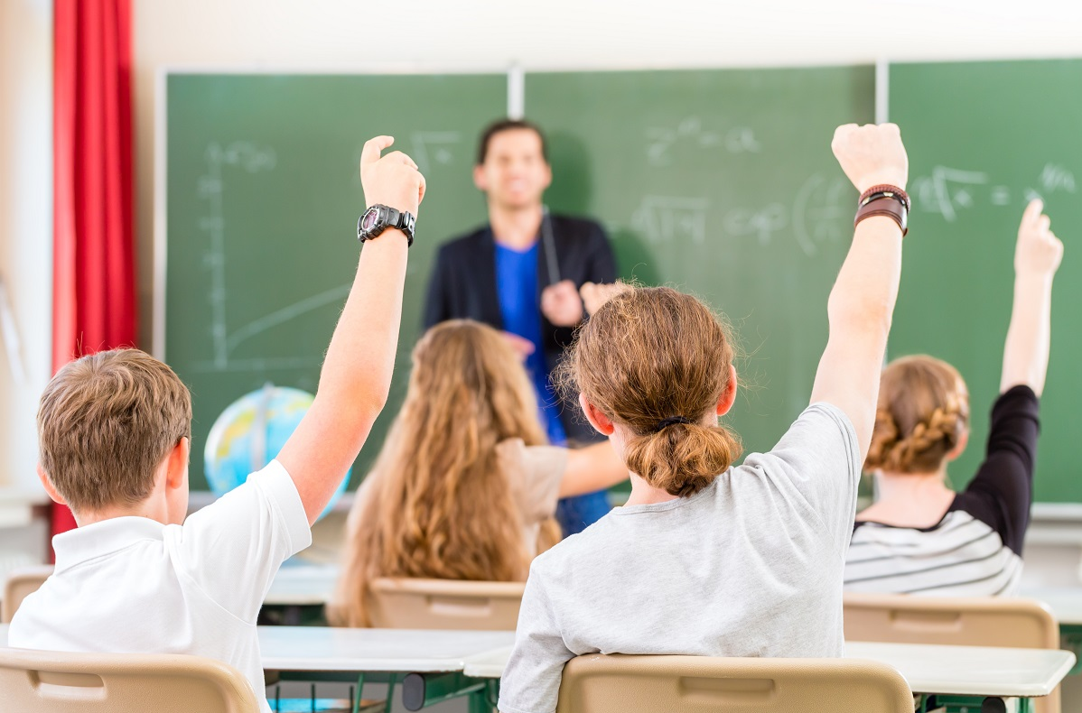 4 students raising hands in class one teacher at blcakboard