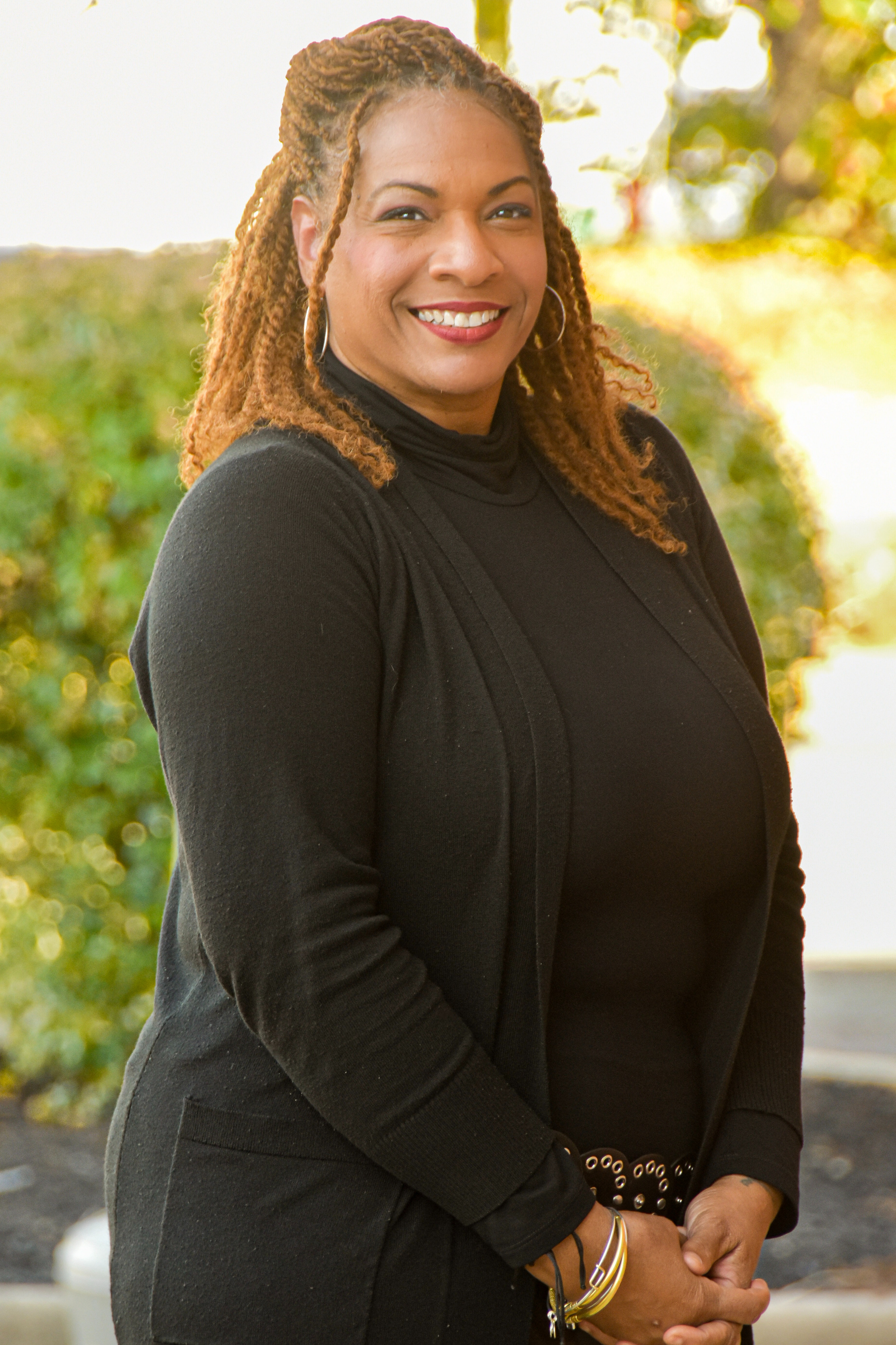 Person with shoulder-length curly hair and a black turtleneck dress stands against a green shrubbery.