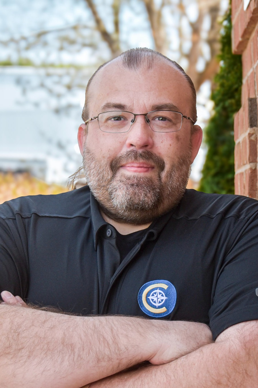 A man with a beard and glasses stands outside, leaning against a brick wall with folded arms.