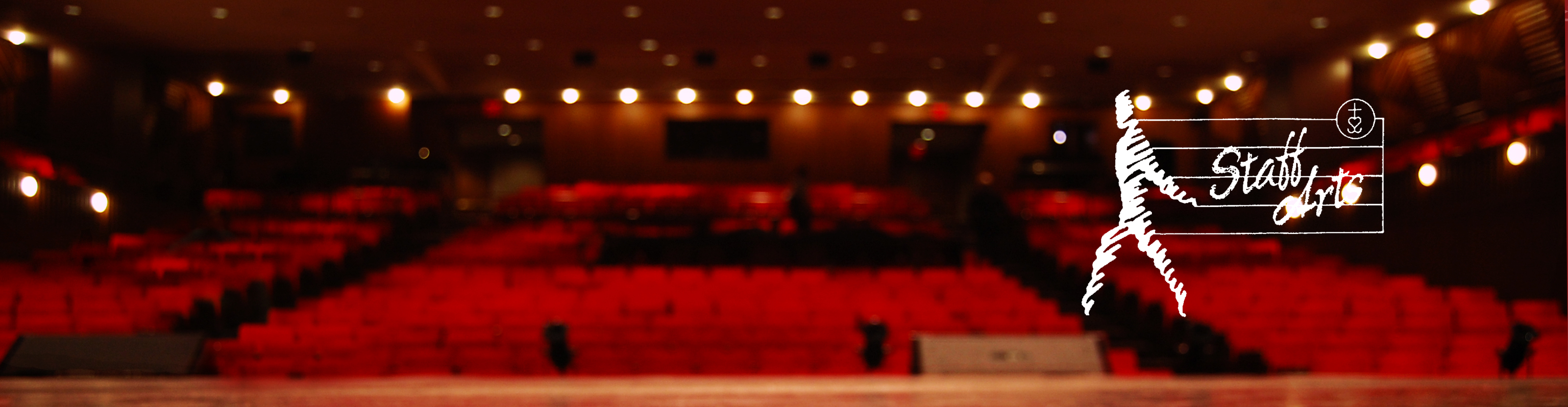 A wide-angle, blurred view of a theater with red seats from the perspective of the stage. The "Staff Arts" logo is overlaid on the right side.