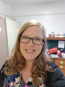 Amy Huffer semi-casual photo with glasses on and a messy bookshelf in the background