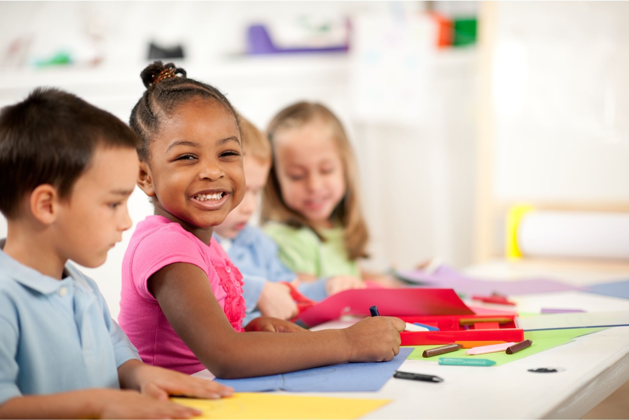 three young children smiling at the camera and drawing on colored paper